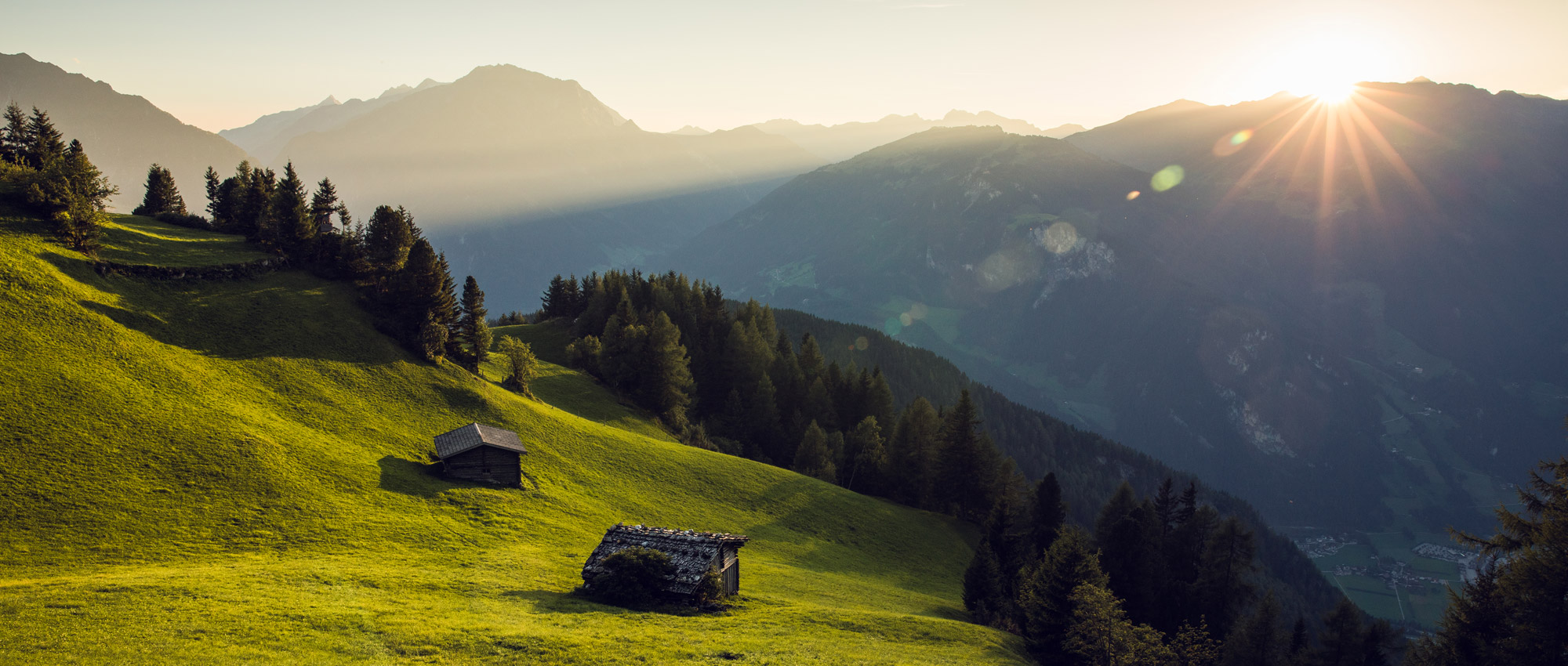 Sonnenaufgang über grünen Berghängen mit zwei Holzhütten, umgeben von Wäldern und Bergen im Hintergrund.